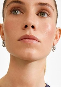 Boucles d'oreilles créoles argentées au design ondulé, finition polie, portées par un mannequin à la peau lisse et maquillage minimal, sur fond neutre.