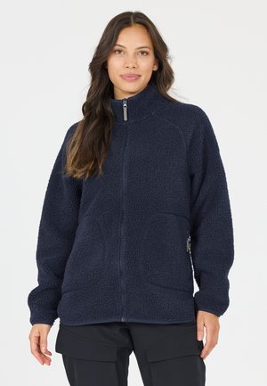 Frau mit langen dunklen Haaren, die eine marineblaue Fleecejacke mit Reißverschluss und schwarze Cargohosen trägt, steht vor einem schlichten, hellen Hintergrund.