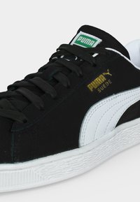 Μαύρο και άσπρο sneaker Puma Suede με μαύρα κορδόνια, λευκή ρίγα, πράσινο λογότυπο στη γλώσσα και χρυσό λογότυπο Puma στο πλάι.