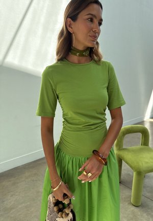 Kvinne iført en tettsittende lys grønn kortermet topp og matchende skjørt, holder en mønstret håndveske, med en grønn stol i bakgrunnen.