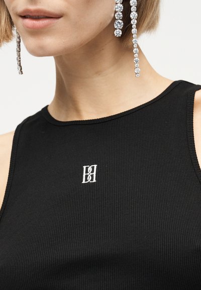 Κοντινό πλάνο ενός ατόμου που φορά μακριά σκουλαρίκια με κρύσταλλα και μια μαύρη αμάνικη ριμπ μπλούζα με ένα μικρό λευκό κεντητό λογότυπο.