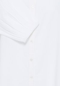 Manche longue blanche avec poignet élastique et boutons blancs visibles le long du plastron avant de la chemise.