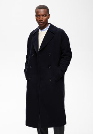 Homme portant un long manteau noir à double boutonnage sur un pull blanc et une chemise bleue à col, debout les mains dans les poches.