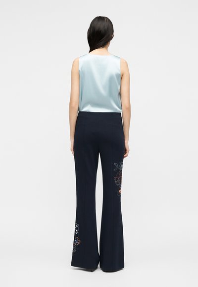 Γυναίκα με μακριά σκούρα μαλλιά που φοράει αμάνικη ανοιχτό μπλε σατέν μπλούζα και μαύρο παντελόνι με φαρδιά πόδια και κεντήματα με λουλούδια στα πόδια, στέκεται με την πλάτη στραμμένη.