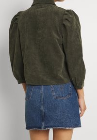 Giacca in corduroy verde oliva con maniche a sbuffo e colletto alto, abbinata a una minigonna in denim con orlo sfrangiato e tasche posteriori.
