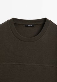 T-shirt a maniche corte verde oliva realizzato in tessuto morbido. Presenta un collo rotondo e un sottile dettaglio di cucitura sul petto. Etichetta del marchio visibile.