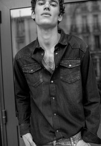 Camicia in denim nero con bottoni, due tasche sul petto, colletto classico e texture leggermente sbiadita. Il modello posa con le mani nelle tasche.