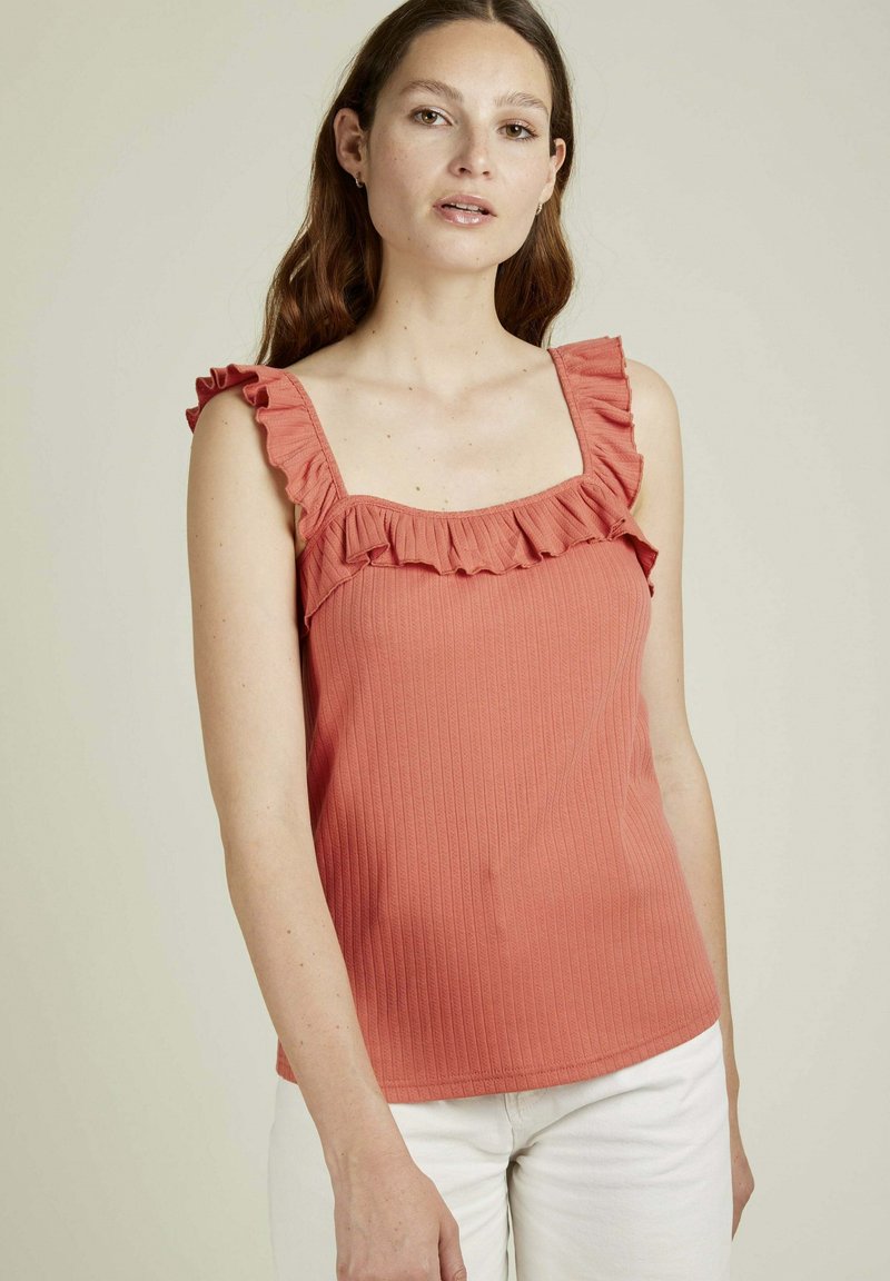 Débardeur côtelé corail avec des accents de volants au niveau du décolleté carré et des bretelles, associé à un pantalon blanc. Tissu lisse et léger.