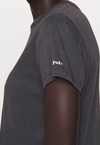 Tmavě šedé tričko s krátkým rukávem vyrobené z bavlny, s kulatým výstřihem a bílým logem na pravém rukávu. Hladká textura.