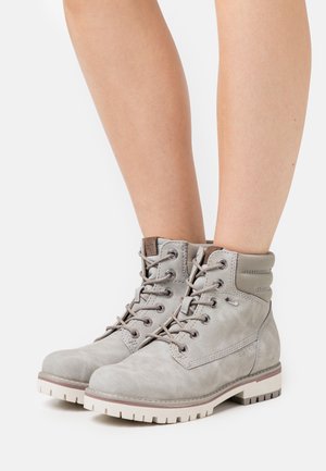 Bottines à lacets gris clair avec col matelassé, œillets en métal, semelle épaisse blanche, portées sur des jambes nues sur un fond blanc.