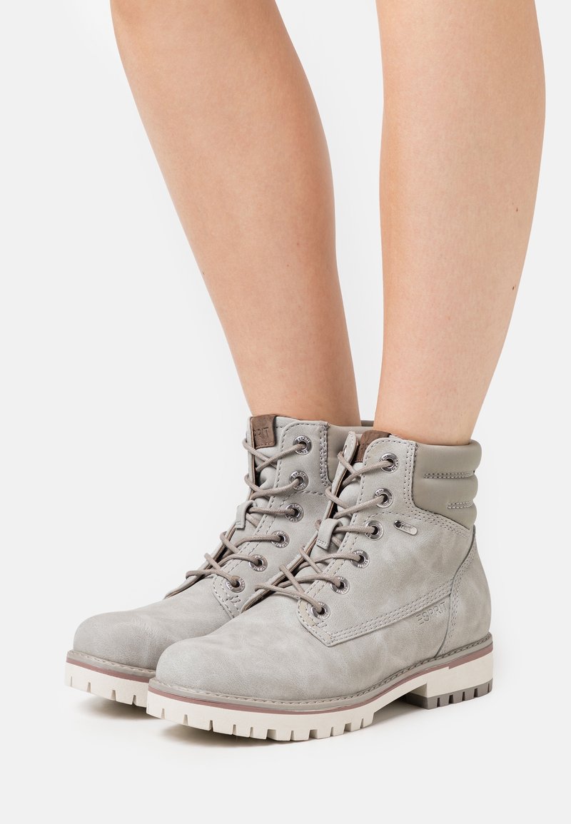 Bottines à lacets gris clair avec col matelassé, œillets en métal, semelle épaisse blanche, portées sur des jambes nues sur un fond blanc.