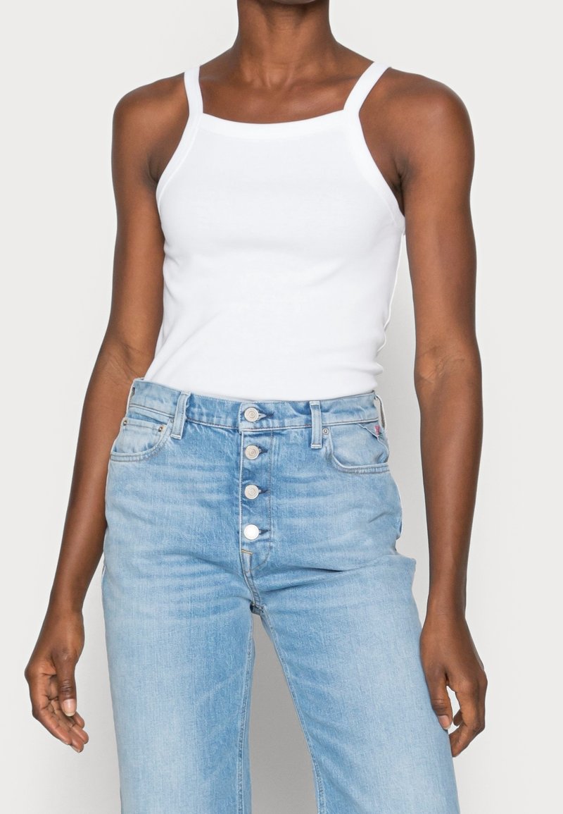 Débardeur blanc à fines bretelles, associé à un jeans en denim bleu clair taille haute avec une fermeture à boutons et un minimum de déchirures.