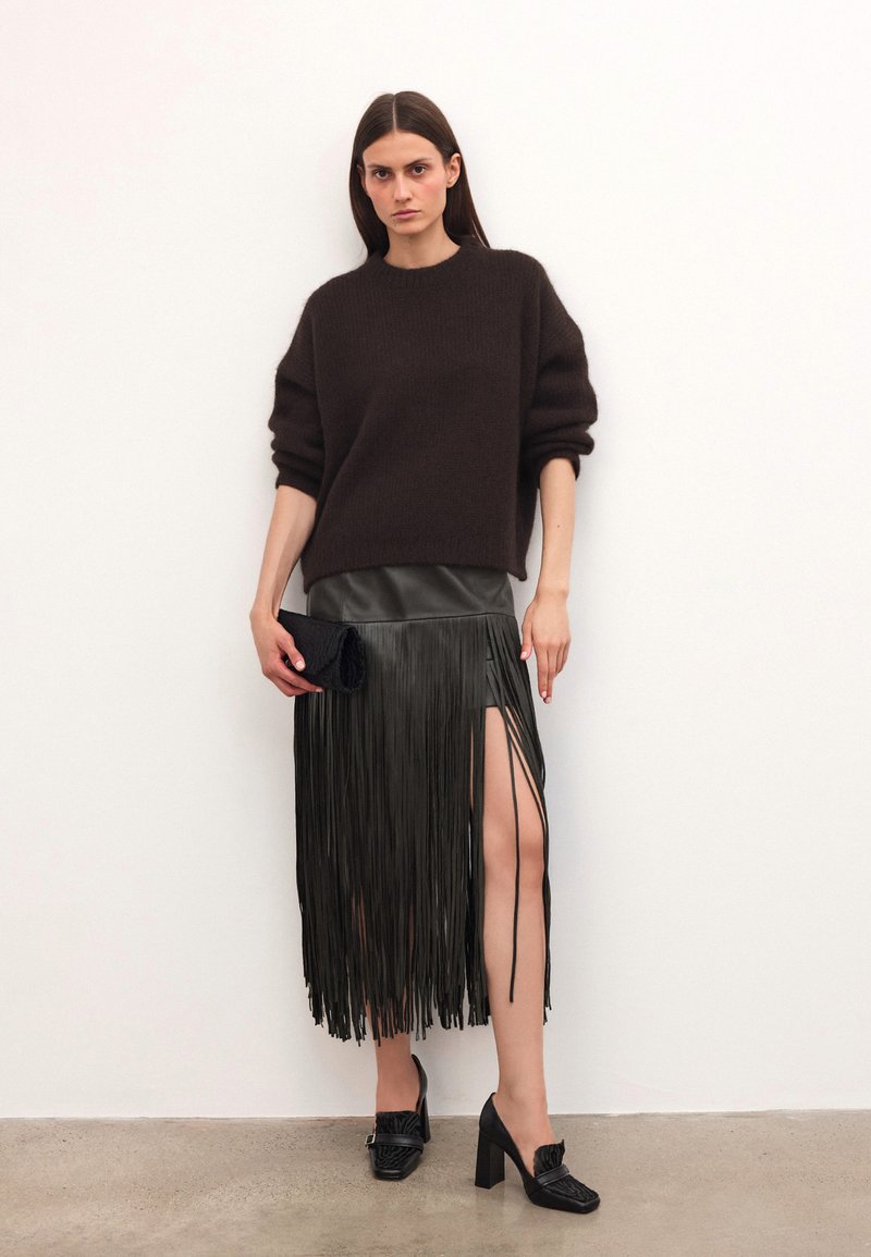 Egy nő barna, oversized pulóvert, fekete bőr frufru szoknyát magas hasítékkal, fekete magassarkú cipőt visel, és egy fekete táskát tart a fehér fal előtt.