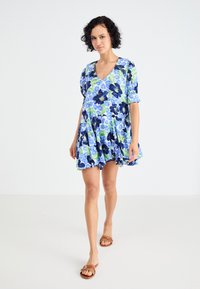 Bloemenjurk met een V-hals, puff mouwen en een uitlopende zoom. De stof is voornamelijk blauw met groene en zwarte bloemmotieven.
