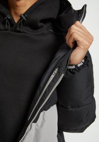 Giubbotto imbottito nero con pannello grigio, chiusura con zip e polsini elasticizzati. Texture liscia con cappuccio regolabile.