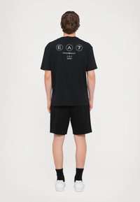 Svart bomull t-skjorte med korte ermer, med et hvitt grafisk logo på baksiden. Kombinert med svarte shorts og hvite joggesko.