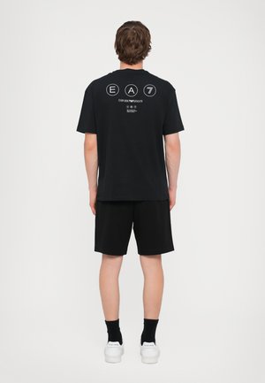 Svart bomull t-skjorte med korte ermer, med et hvitt grafisk logo på baksiden. Kombinert med svarte shorts og hvite joggesko.