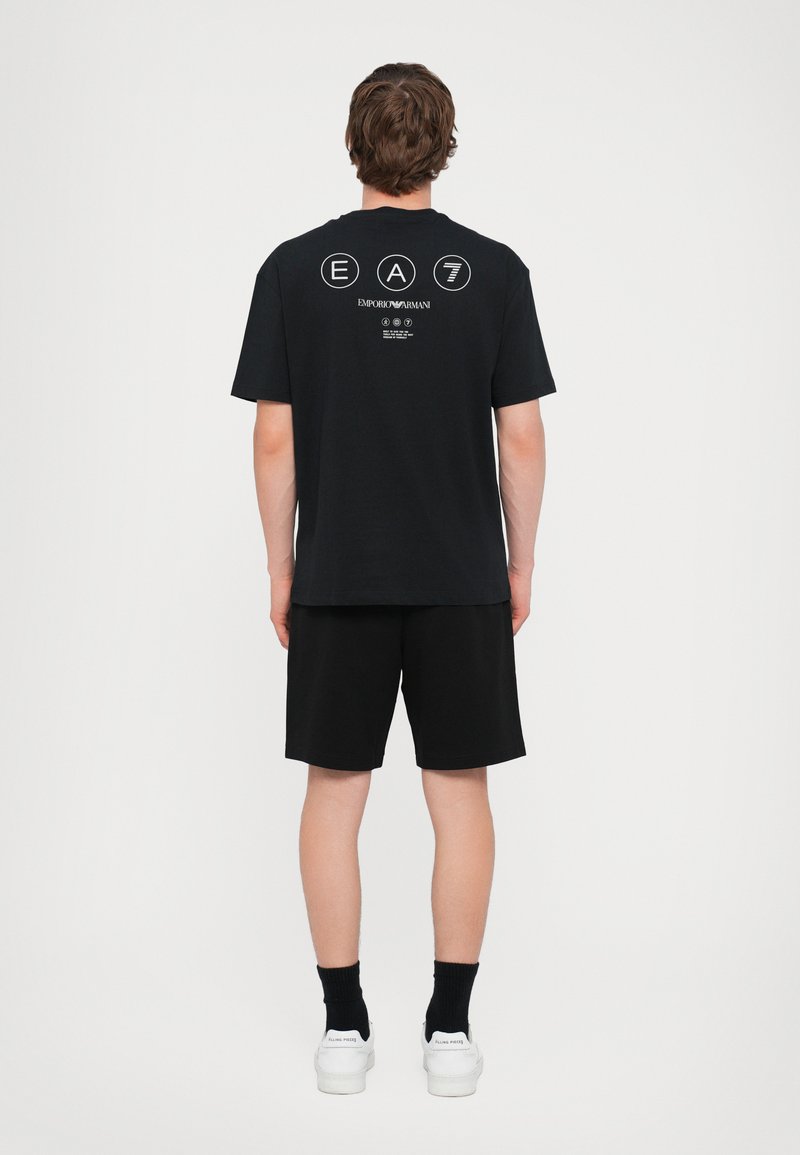 Svart bomull t-skjorte med korte ermer, med et hvitt grafisk logo på baksiden. Kombinert med svarte shorts og hvite joggesko.