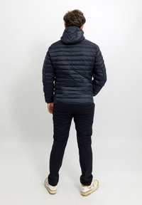 Giacca imbottita blu navy con cappuccio, caratterizzata da un motivo trapuntato. Indossata con pantaloni scuri e sneakers di colore chiaro, mostrata di schiena.