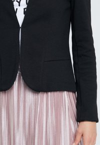 Czarna dopasowana marynarka o gładkiej fakturze, z otwartym przodem i dwoma bocznymi kieszeniami, w połączeniu z plisowaną różową metaliczną spódnicą.