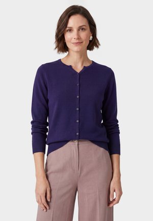 Donna con un cardigan viola abbottonato e pantaloni color marrone chiaro a vita alta, in piedi davanti a uno sfondo chiaro uniforme.