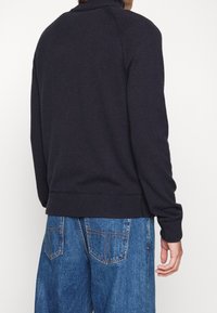 Mörkblå stickad tröja med hög krage, ribbad mudd och fåll. Bärs med blå denimbyxor som har bakfickor och synlig sömnad.