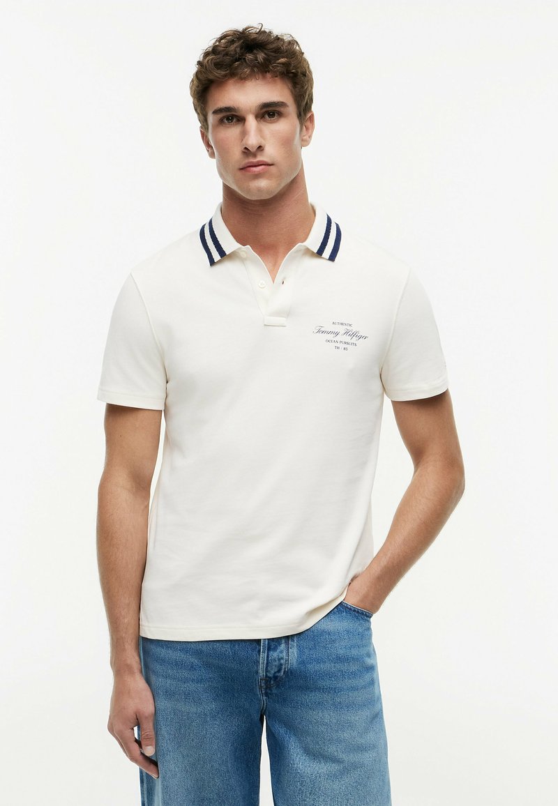 Jeune homme aux cheveux bouclés portant un polo blanc avec un col rayé bleu marine et un jean bleu, debout avec une main dans la poche.