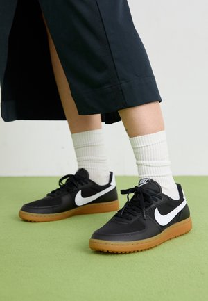 Nogi w białych skarpetach typu crew i czarnych butach Nike z białym znakiem i gumowymi podeszwami, stojące na jasnozielonej podłodze.