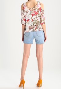 Žena oblečená v bílé květinové blůze s odhalenými rameny, světle modrých denimových šortkách a oranžových lodičkách, stojící s otočením zády.