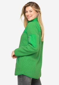 Zielony polarowy sweter z wysokim kołnierzem, pikowanymi zielonymi akcentami na ramionach oraz kieszenią z zamkiem błyskawicznym na rękawie, wykonany z miękkiego, teksturowanego materiału.