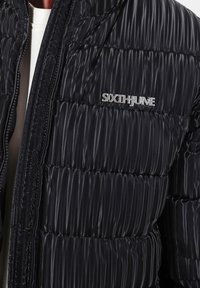 Zwarte pufferjack met horizontaal gewatteerd ontwerp, voorzien van een zilveren logo op de borst, glanzende textuur en ritssluiting aan de voorkant.