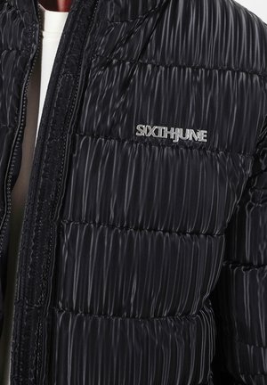 Zwarte pufferjack met horizontaal gewatteerd ontwerp, voorzien van een zilveren logo op de borst, glanzende textuur en ritssluiting aan de voorkant.