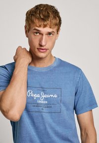 Zila kokvilnas T-krekls ar īsām piedurknēm, ar balto "Pepe Jeans London" grafiku priekšpusē, apaļu kakla izgriezumu un mīkstu tekstūru.