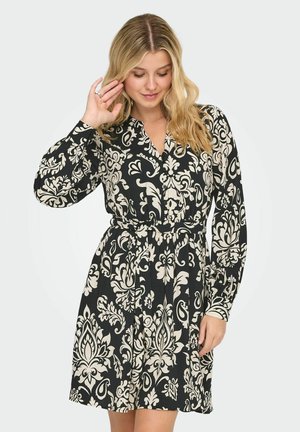 Vrouw met lang blond haar, gekleed in een zwart-beige jurk met lange mouwen en patroon, poseert met één hand bij haar gezicht terwijl ze naar beneden kijkt.