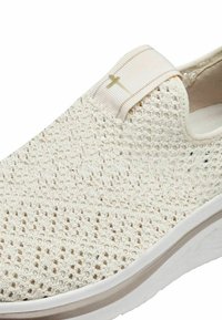 Strickschuh in Creme mit strukturiertem Obermaterial, elastischem Band am Knöchel und einer weißen Zwischensohle, die durch einen subtilen beigen Streifen akzentuiert ist.