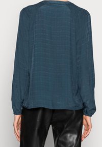 Blusa leggera color teal con tessuto testurizzato, maniche lunghe e polsini elasticizzati. Presenta un collo rotondo e una vestibilità comoda, abbinata a pantaloni neri.