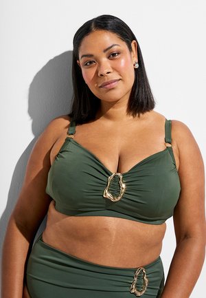 Kobieta o średnim odcieniu skóry i prostych czarnych włosach nosi oliwkowozielony strój kąpielowy z złotymi ozdobnymi zapięciami, stojąc na tle gładkiej białej ściany.