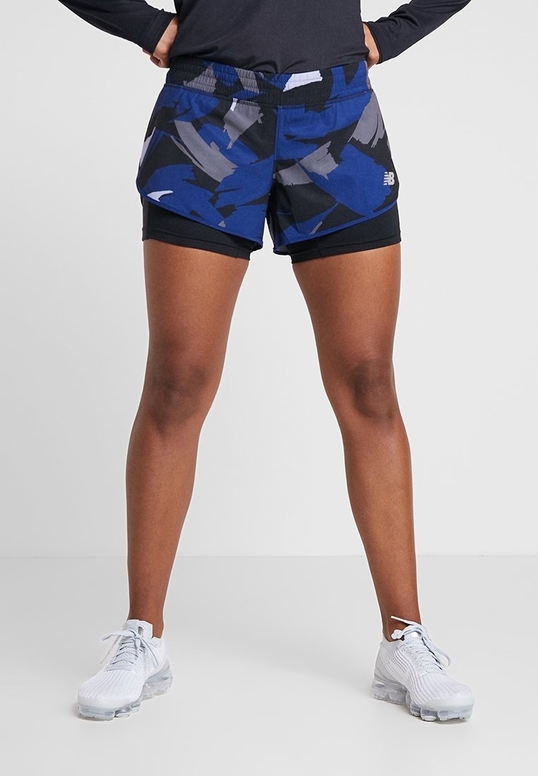 Osoba stojící s rukama v bok, oblečená v černých a modrých vzorovaných sportovních šortkách a bílých běžeckých botách na jednobarevném pozadí.