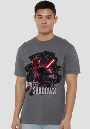 Young man wearing a gray T-shirt featuring a red and black graphic of Darth Maul holding a red lightsaber with text "From the Shadows."