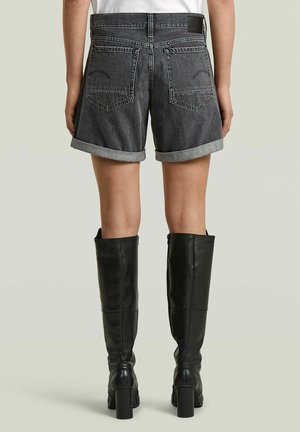 Unterer Torso mit hochgekrempelten schwarzen Jeansshorts und schwarzen kniehohen Stiefeln mit Absatz, stehend vor hellem einfarbigen Hintergrund.