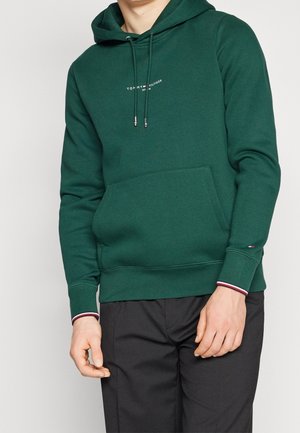 Osoba ubrana w ciemnozieloną bluzę z kapturem Tommy Hilfiger z przednią kieszenią i sznurkami, w połączeniu z czarnymi spodniami, na jednolitym tle.