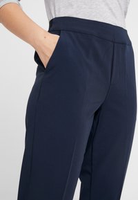 Pantalon bleu marine sur mesure avec une texture lisse, poches latérales et une taille ajustée, conçu avec un minimum de coutures pour un look épuré.