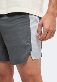 Homme portant un short de sport gris bicolore avec un petit symbole de copyright sur la jambe gauche près d'une fente latérale.