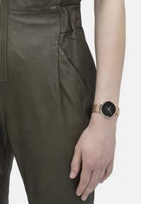 Gouden horloge met een zwarte wijzerplaat, ronde klok, en een geschakelde metalen band, gedragen op een hand tegen olijfgroen leren kleding.