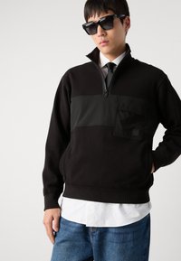 Felpa nera con colletto a zip e tasca frontale, caratterizzata da un design testurizzato. Abbinata a una camicia bianca e jeans blu.