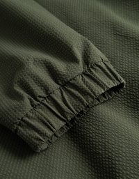 Textúrovaná tmavozelená tkanina s ryhovaným vzorom, so zhromaždenou elastickou manžetou pre pohodlné nosenie. Hladký povrch s jemným leskom.