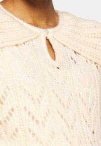 Kabelstickad krämfärgad tröja med hög krage, nyckelhålsdetalj och dekorativ knapp vid halsen, som visar upp ett mjukt, texturerat tyg.
