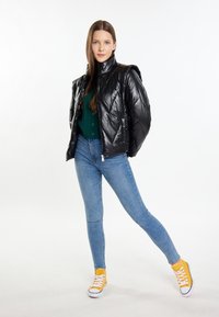 Fekete puffdzseki magas gallérral, zöld trikó pulóver, slim fit kék farmer, és sárga vászon sportcipő fehér talppal.