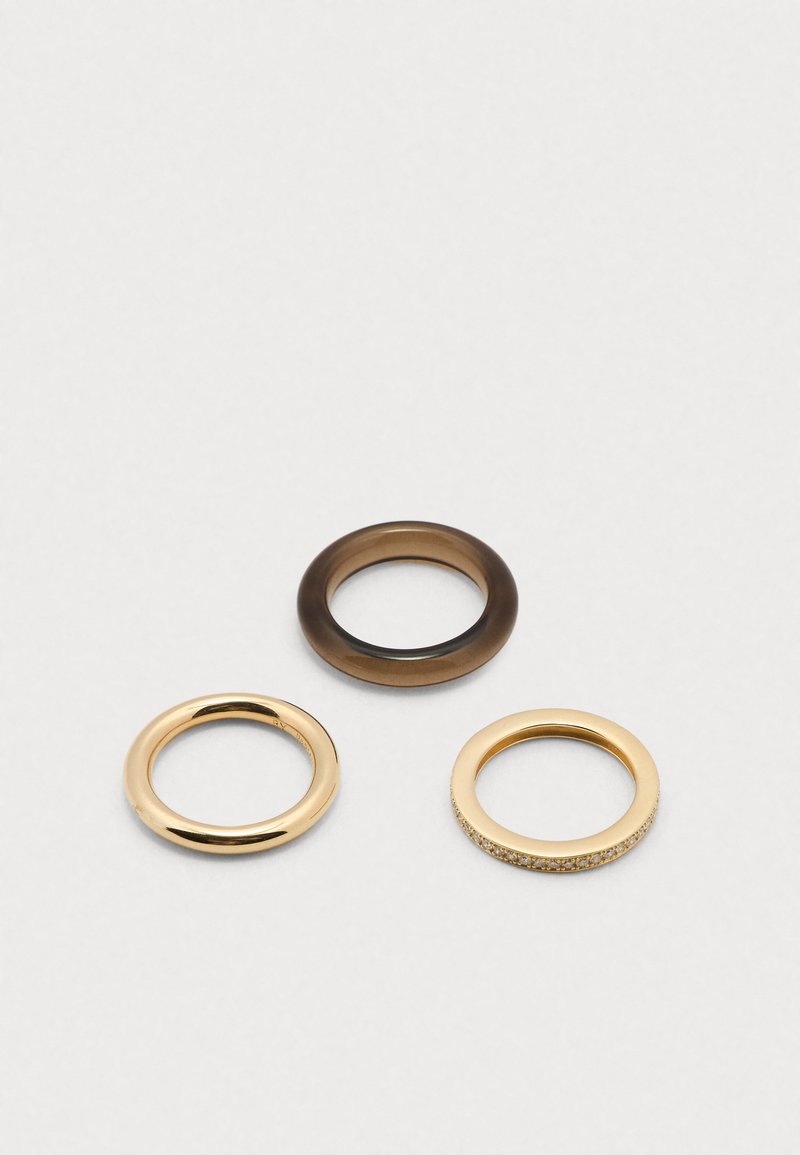 Három gyűrű: egy masszív barna gyanta gyűrű, egy sima arany fém gyűrű, valamint egy arany gyűrű, amelynek texturált felületét apró kristályok díszítik.