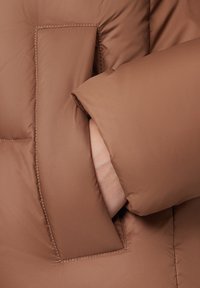 Rękaw brązowej kurtki puchowej z dużą, prostokątną kieszenią. Gładki, lekko błyszczący materiał z miękką podszewką widoczną wewnątrz kieszeni.